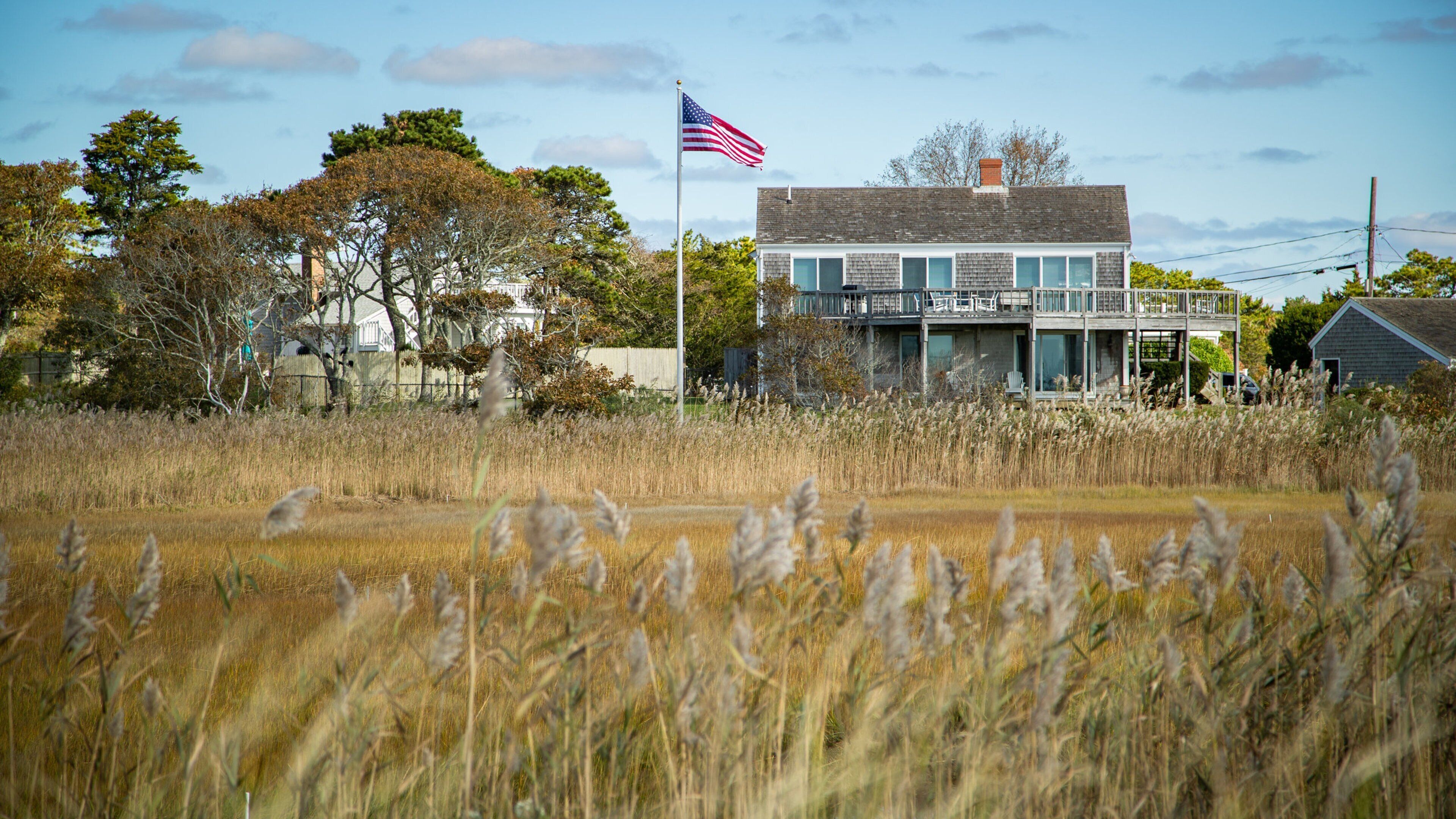 A two-story house with a flagpole and American flag, surrounded by tall grass and trees.