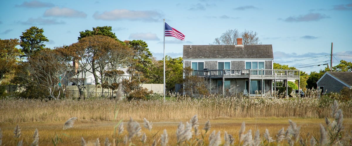 A two-story house with a flagpole and American flag, surrounded by tall grass and trees.