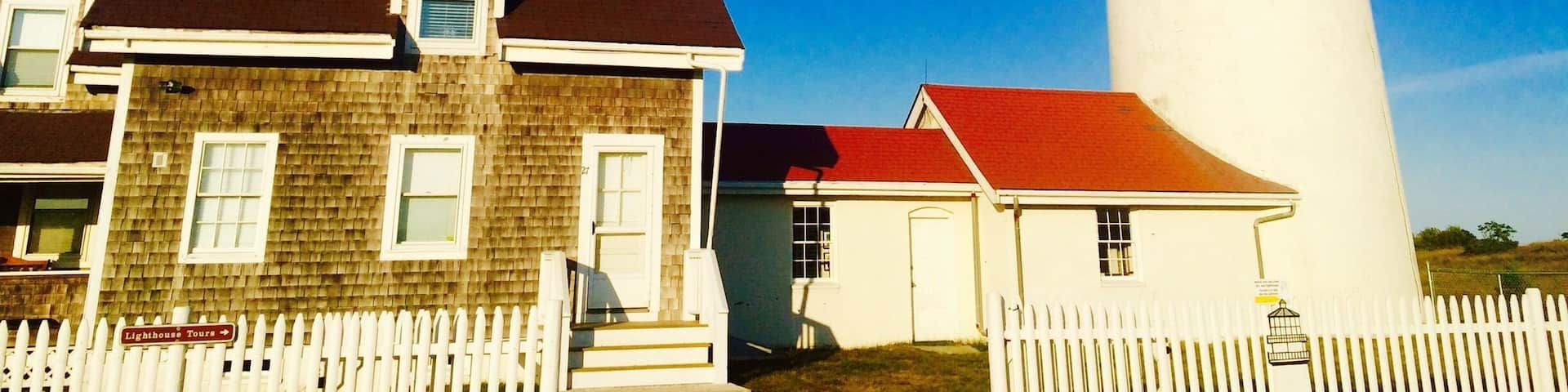 A lighthouse with a white tower and black top, a wooden house with a red roof, and a white picket fence.