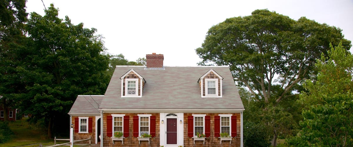 A two-story brick house with a red door and shutters, surrounded by a wooden fence and trees.