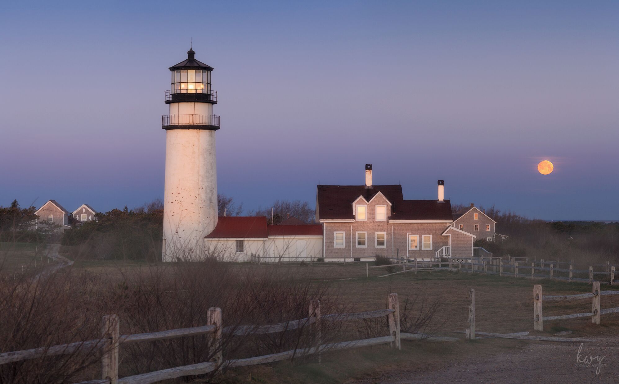 A lighthouse, a brick house, and a wooden fence in a rural setting.