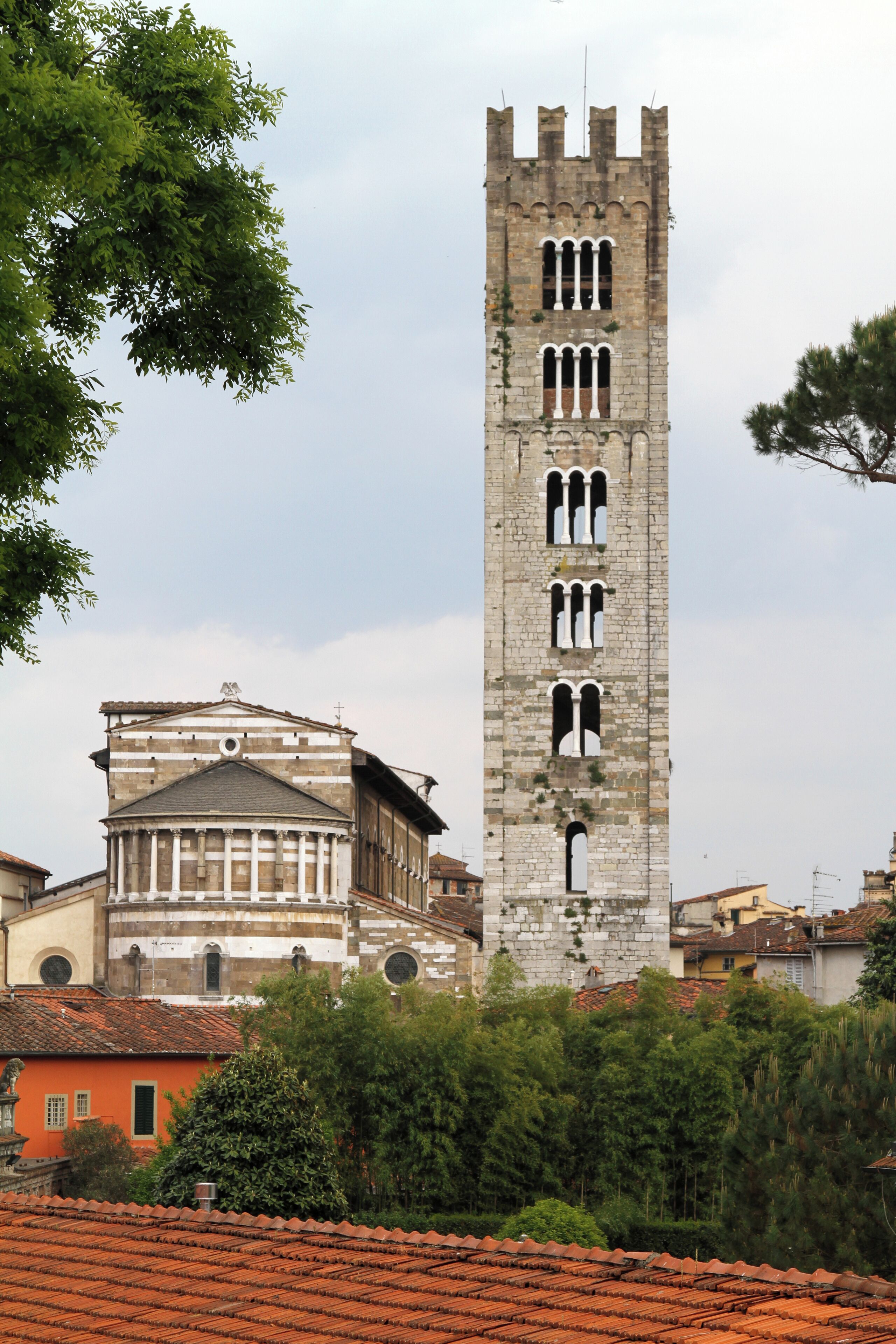 A tall stone tower with arched windows and a bell tower, surrounded by trees and buildings with red-tiled roofs.