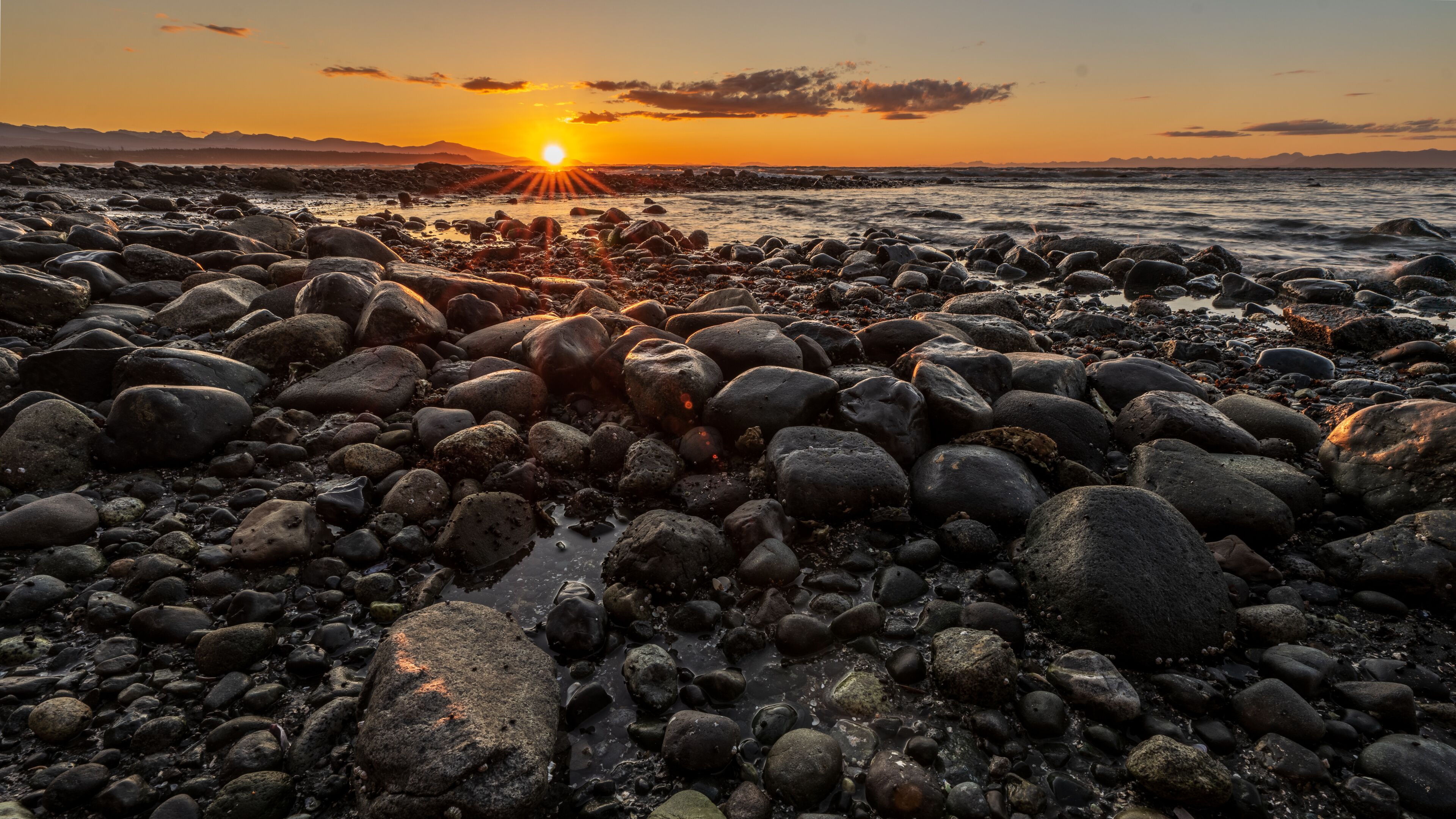 A rocky shoreline at sunset with the sun low on the horizon.