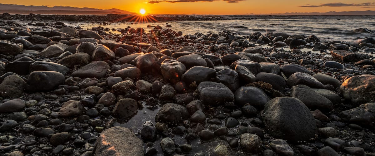 A rocky shoreline at sunset with the sun low on the horizon.