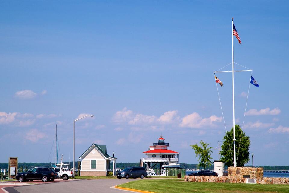 A lighthouse, a small building, a flagpole with flags, a one-way sign, and a body of water.