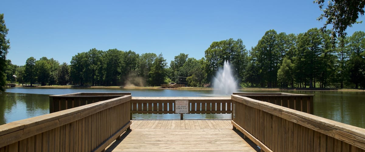 A wooden pier leading to a lake with a fountain in the center.