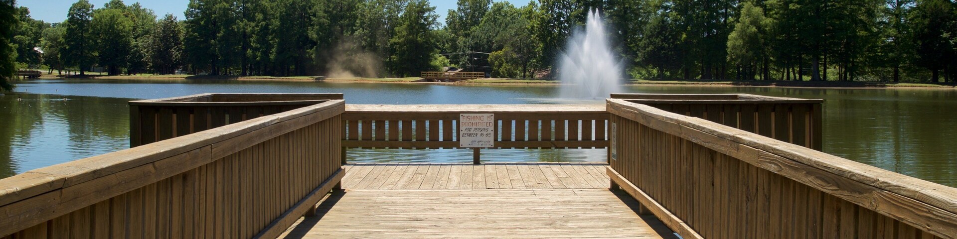 A wooden pier leading to a lake with a fountain in the center.