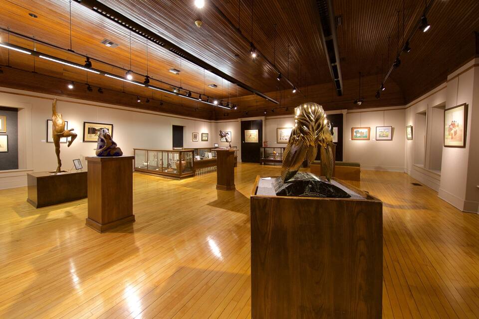 Uma galeria de arte com pisos e teto de madeira, exibindo esculturas e obras de arte emolduradas.