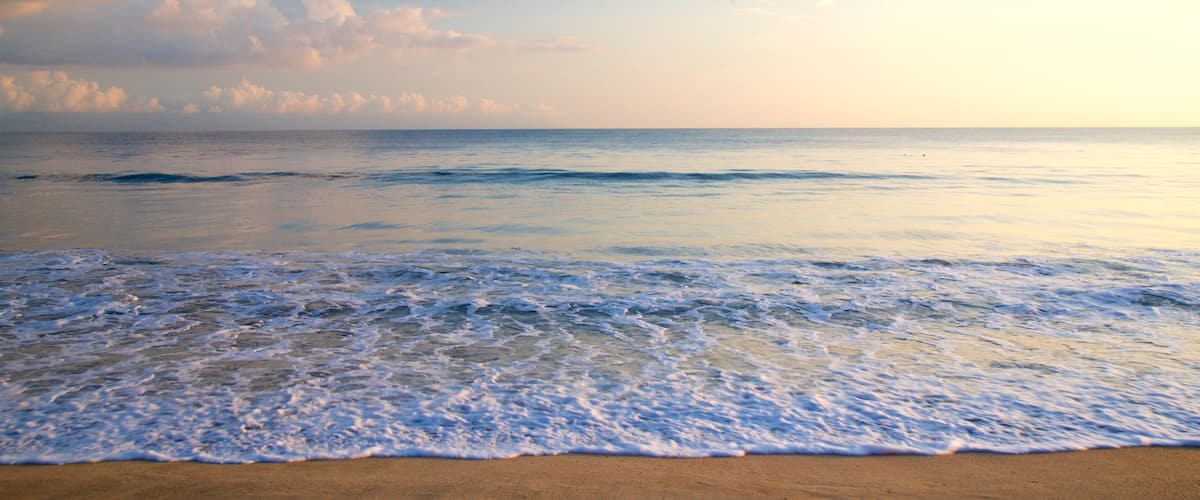 A beach with waves gently lapping onto the shore, a clear sky with scattered clouds, and a calm sea.