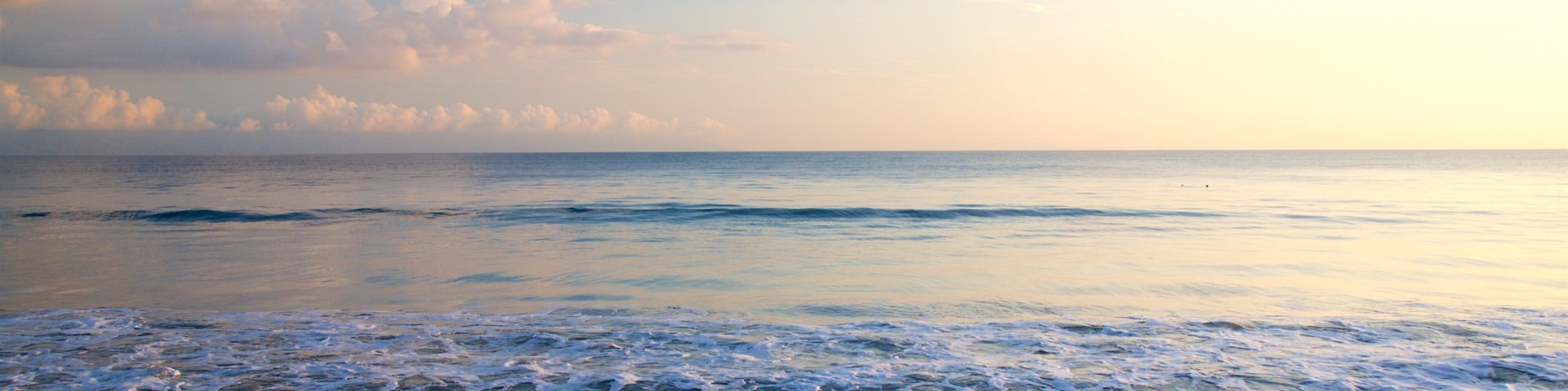 A beach with waves gently lapping onto the shore, a clear sky with scattered clouds, and a calm sea.