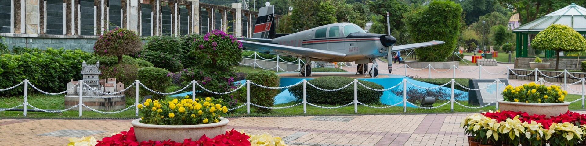 Un avión militar exhibido, rodeado de plantas en macetas y una cerca de malla metálica.