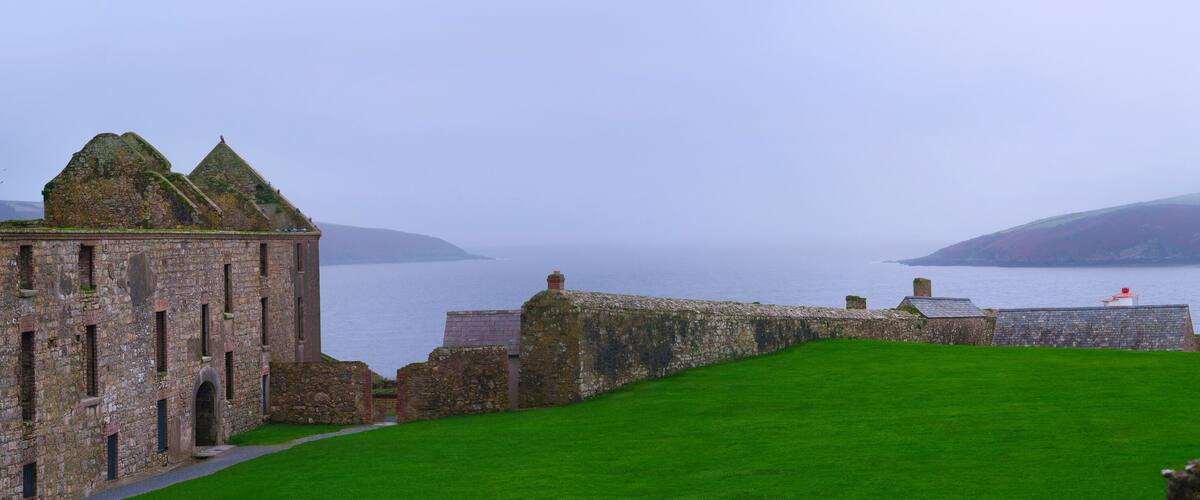 A historic stone building with a grassy courtyard and a view of the sea.