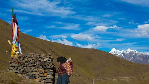 Un paisaje montañoso con cielo azul despejado, una construcción de piedra y una persona con un chal colorido.