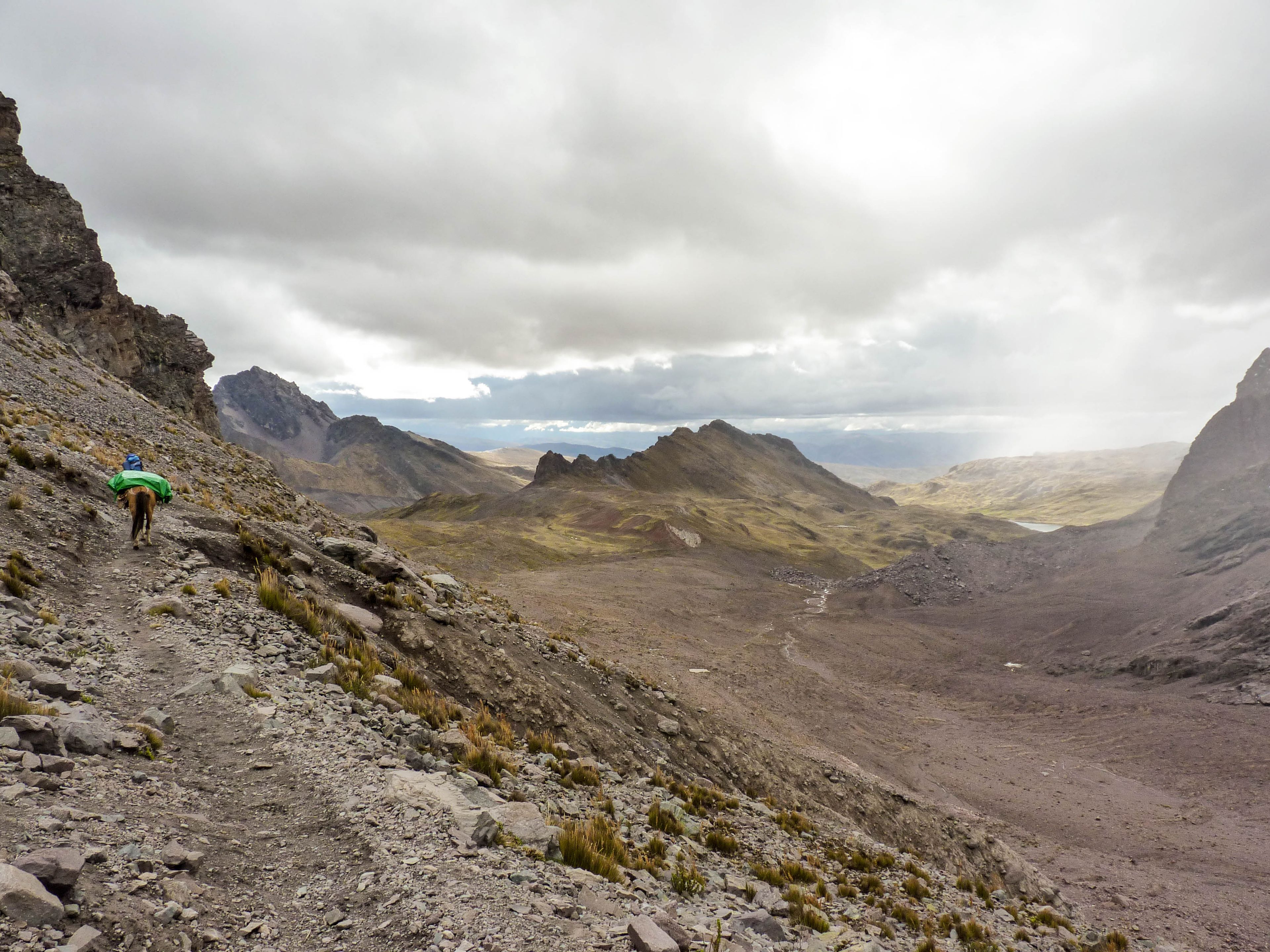 A rugged mountain trail with a hiker and a horse, surrounded by rocky terrain and distant peaks under a cloudy sky.