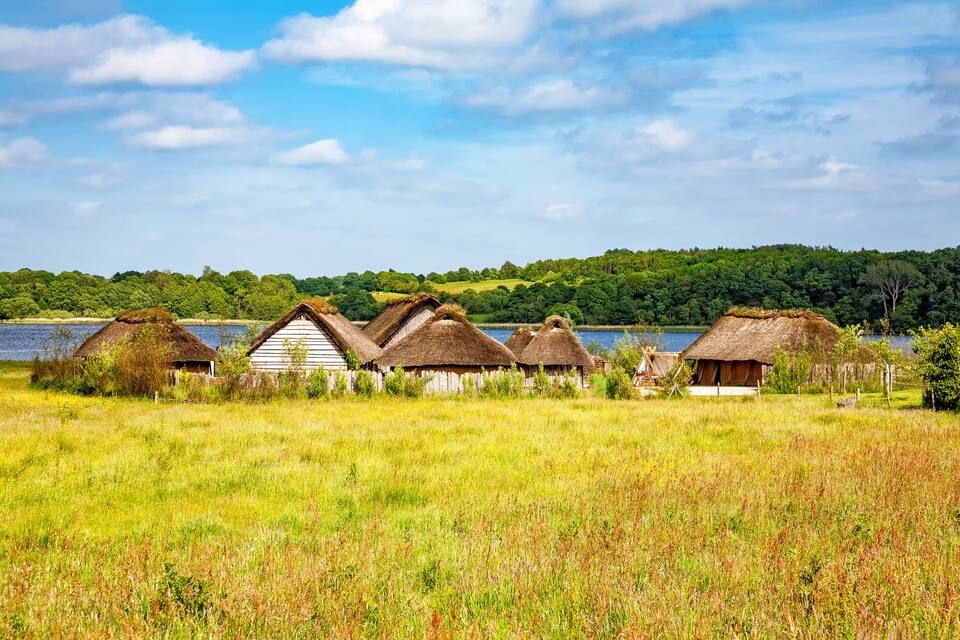 Reetgedeckte Hütten in einem grasbewachsen Feld, mit einem See und einem Wald im Hintergrund.