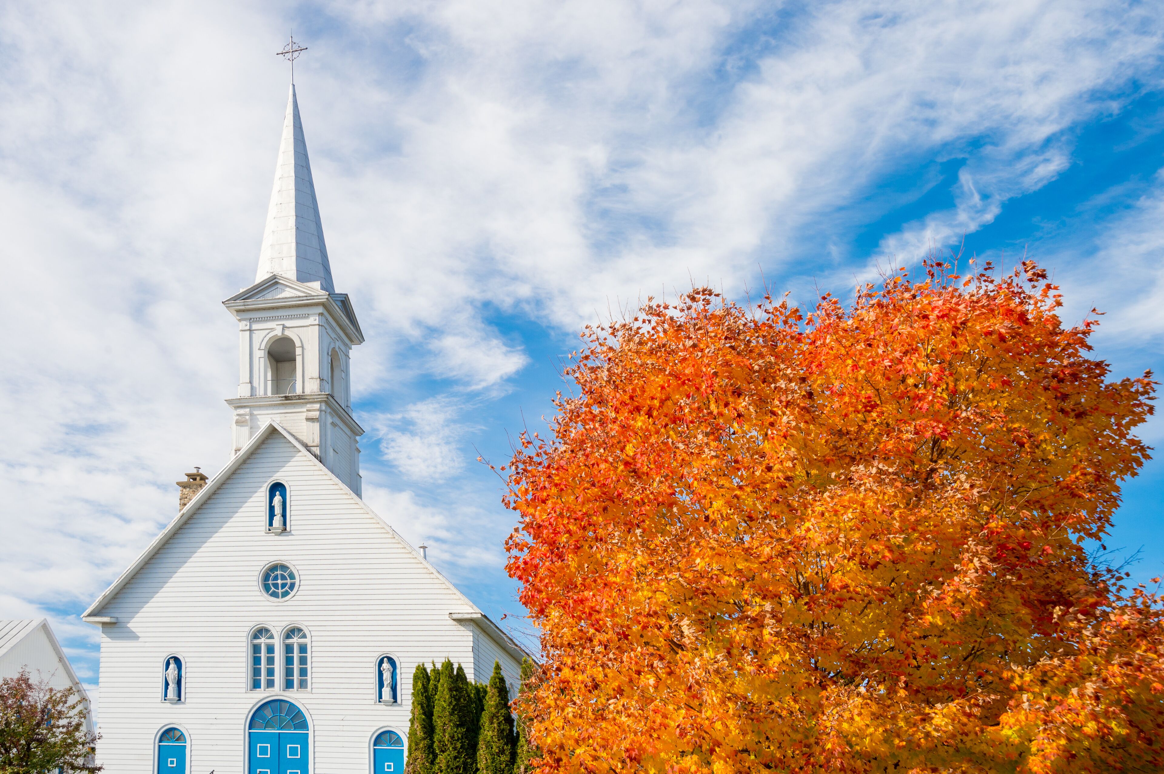 A white church with a tall steeple and blue doors, surrounded by autumn foliage.
