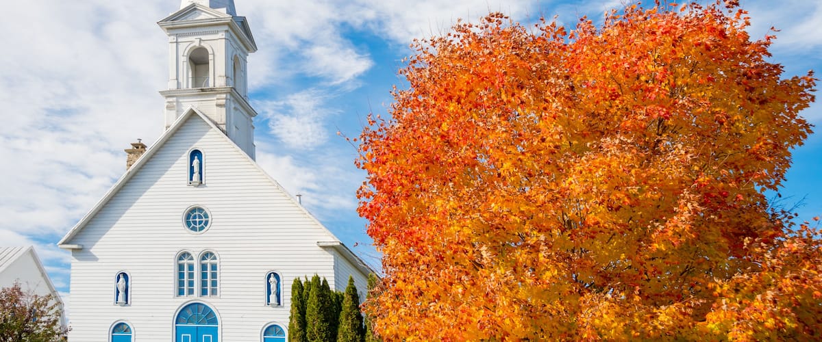 A white church with a tall steeple and blue doors, surrounded by autumn foliage.