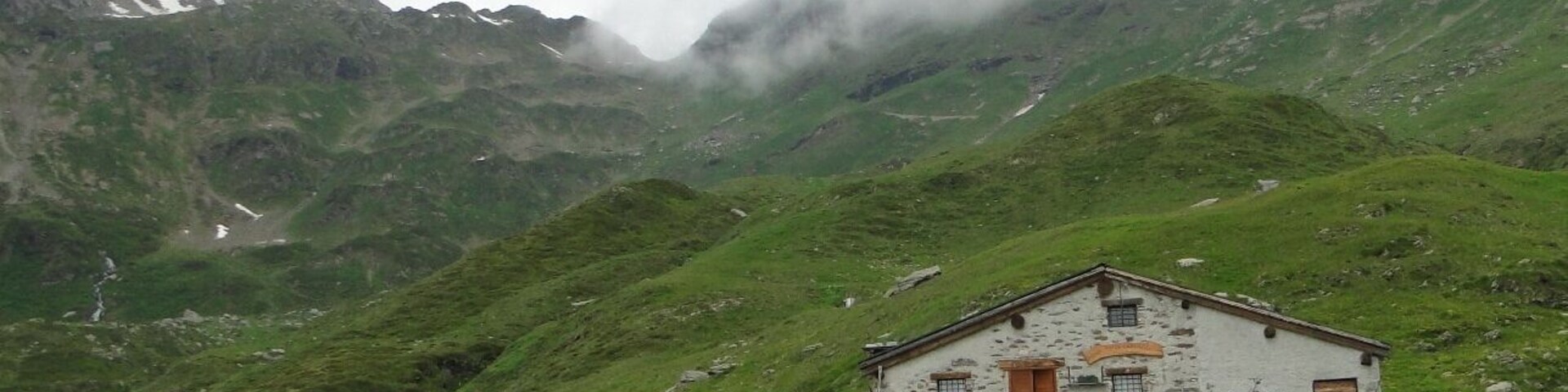 Una casa de piedra sobre una ladera cubierta de hierba, con montañas y nubes al fondo.