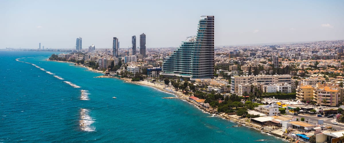 Aerial view of Limassol, Cyprus, featuring modern high rise buildings, a curved structure, turquoise sea, sandy beach, and wave breakers.