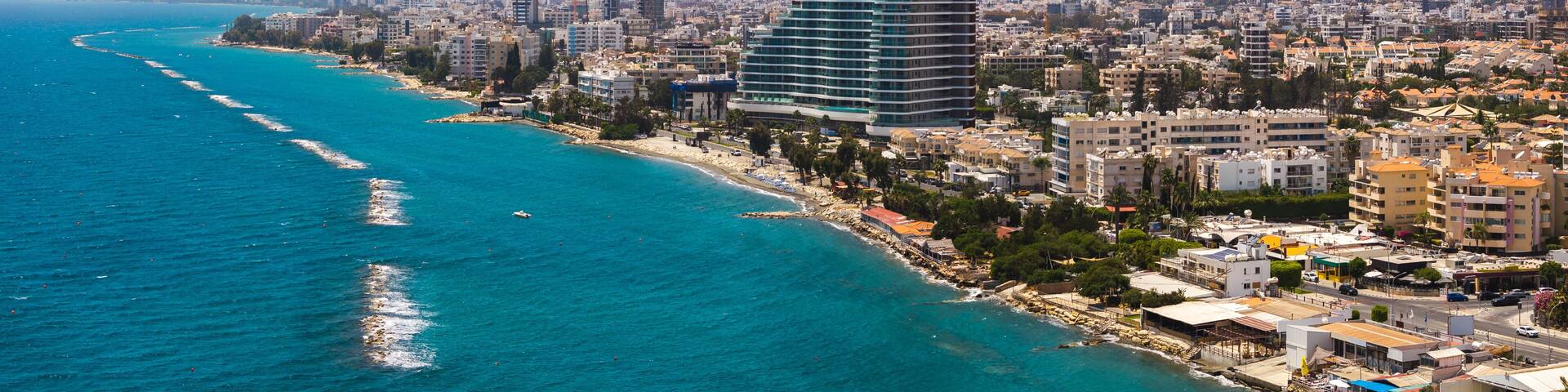 Aerial view of Limassol, Cyprus, featuring modern high rise buildings, a curved structure, turquoise sea, sandy beach, and wave breakers.