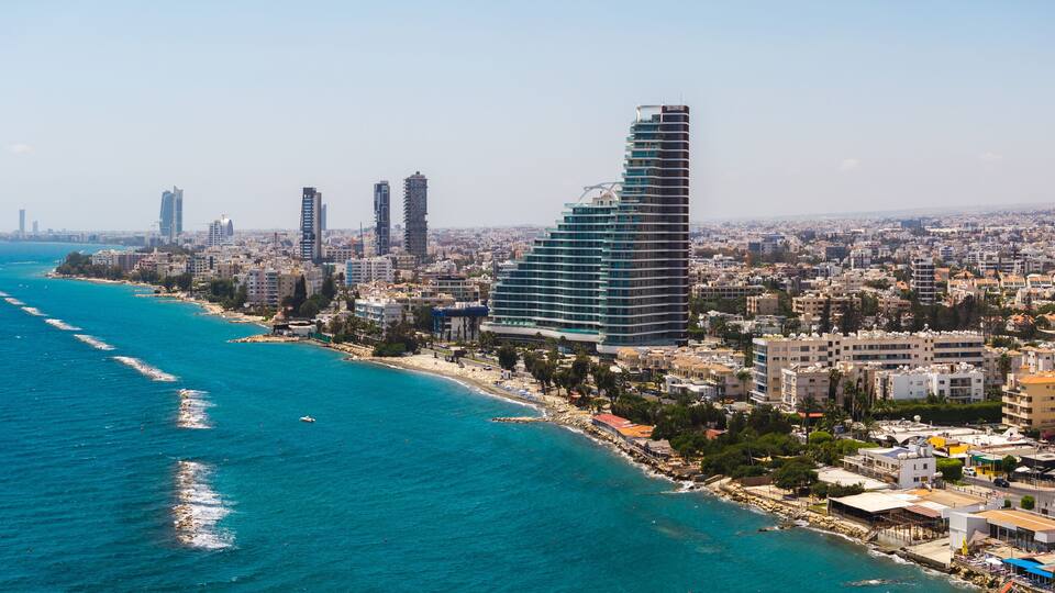 Aerial view of Limassol, Cyprus, featuring modern high rise buildings, a curved structure, turquoise sea, sandy beach, and wave breakers.