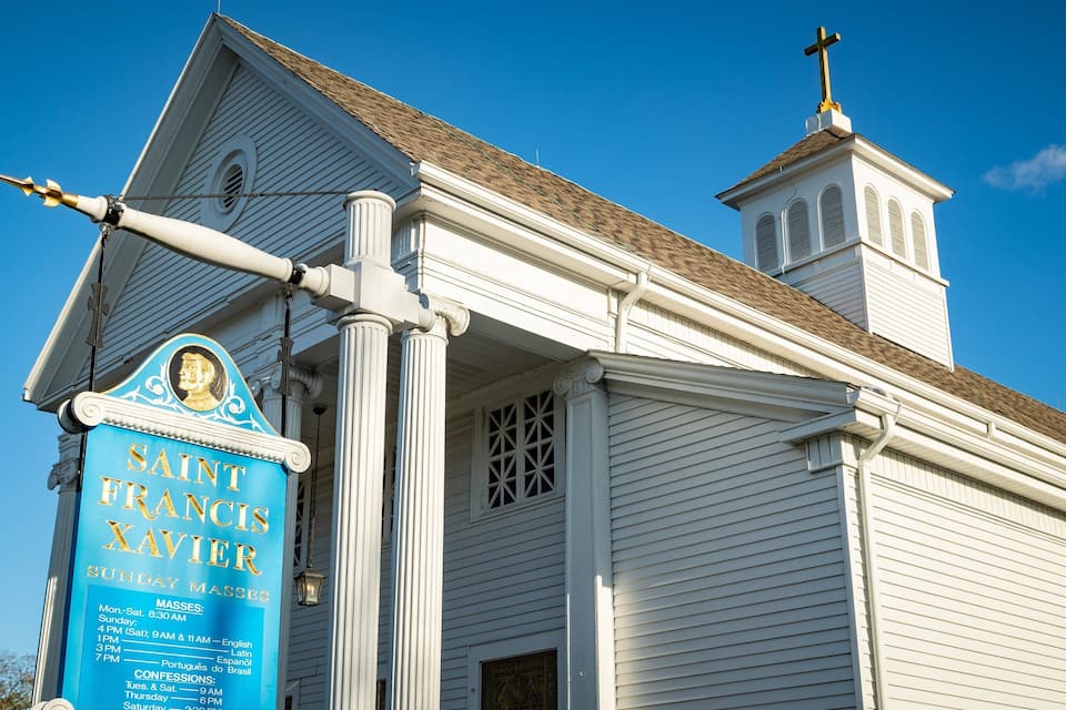A white church with a steeple and a sign for Saint Francis Xavier Sunday masses.