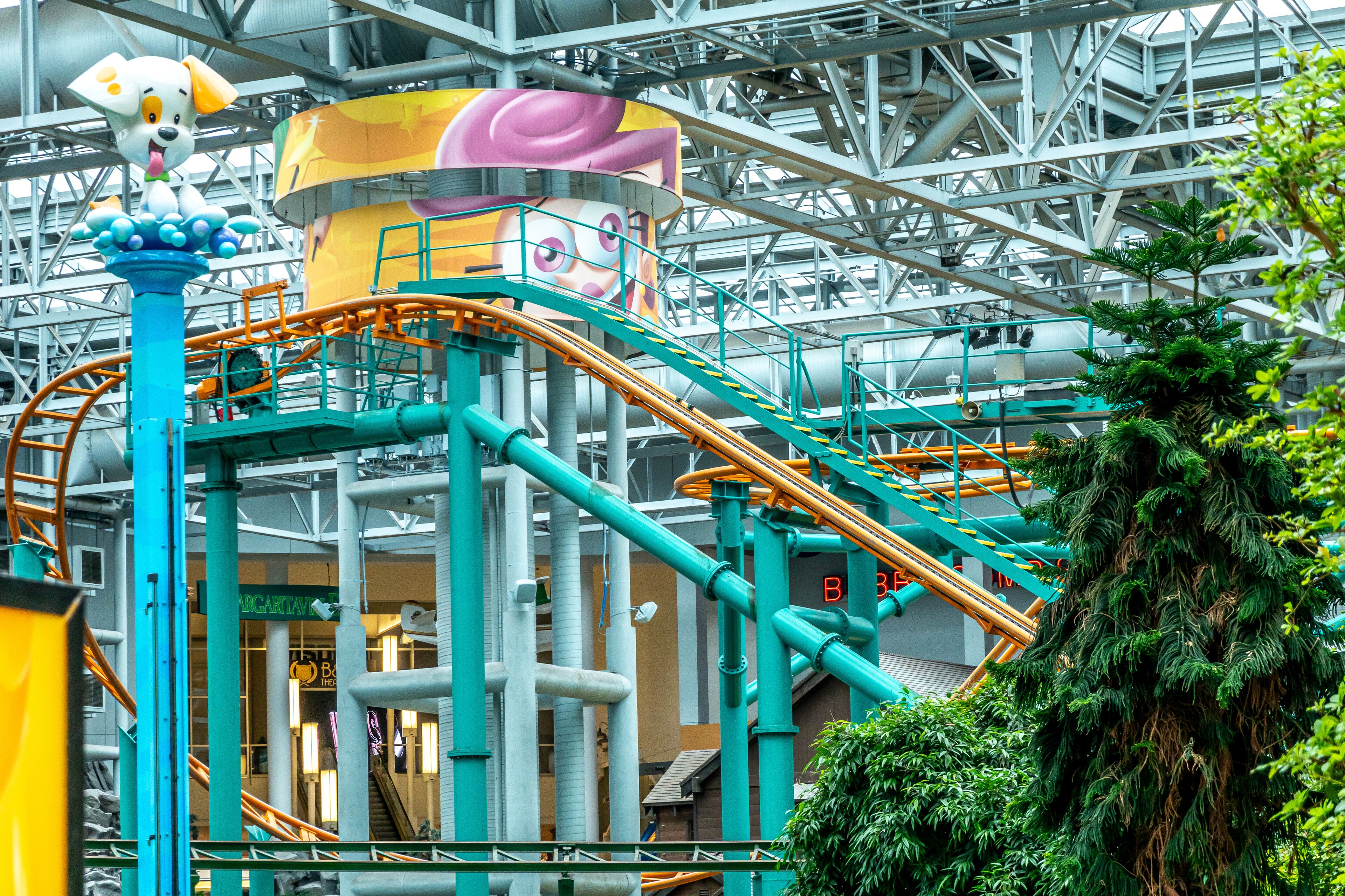 A roller coaster with a large mascot figure, a green tree, and industrial structures.
