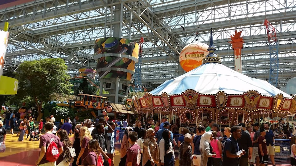 A crowded indoor amusement park with a large Nickelodeon Universe sign, a carousel, and a roller coaster.