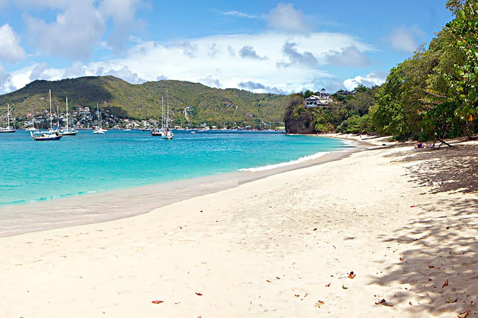 Une plage bordée d’une eau turquoise et limpide, avec des bateaux et un paysage montagneux en arrière-plan.