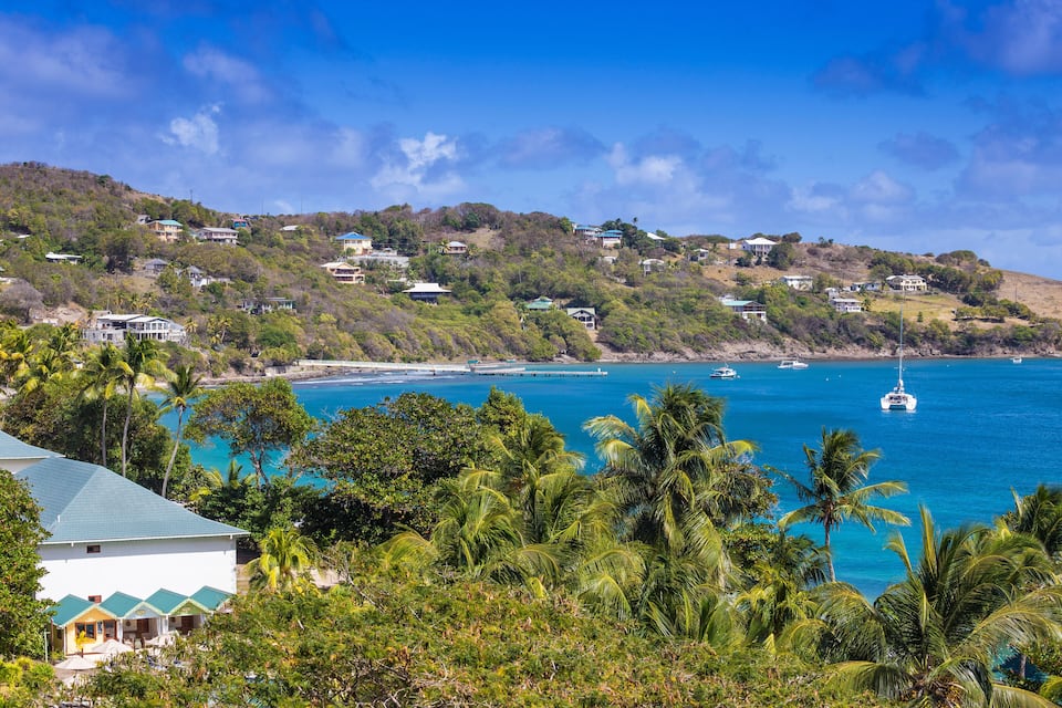 Un paysage côtier avec des maisons sur une colline, une mer bleue et limpide, et un voilier.