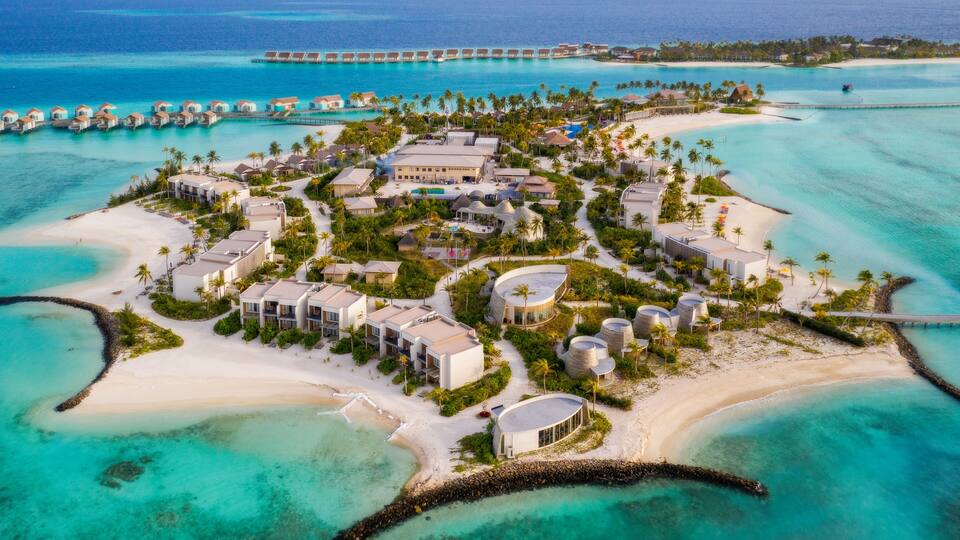 An aerial view of a resort with overwater bungalows, a beach, and clear turquoise waters.