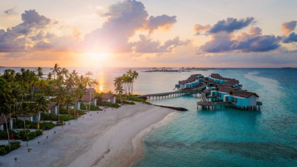 A beach resort with overwater bungalows, palm trees, and a sunset over the ocean.