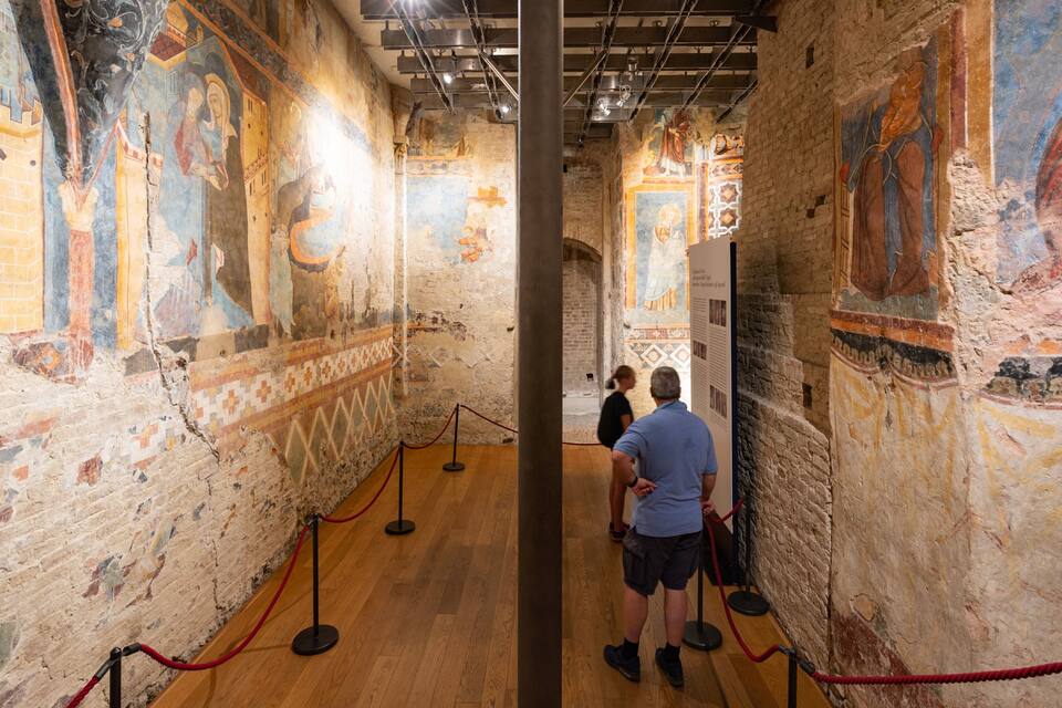 Interior com afrescos antigos nas paredes, soalho de madeira e uma barreira de corda vermelha.