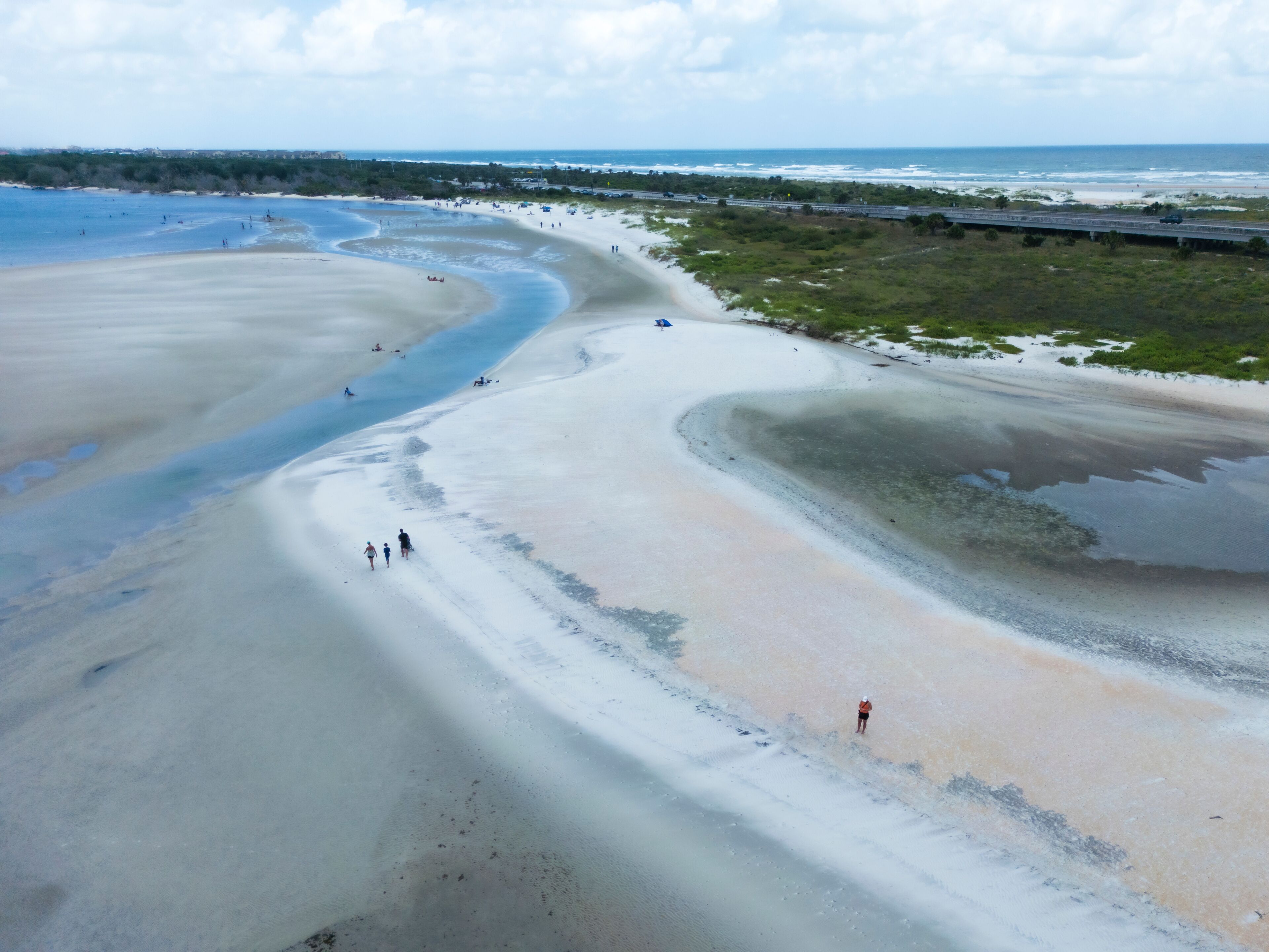 Paisagem costeira com uma extensa praia de areia, ondas suaves e uma rodovia distante.