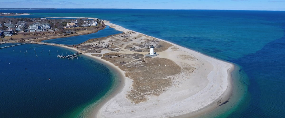 An aerial view of a coastal area with a lighthouse, a sandy beach, and a marina.