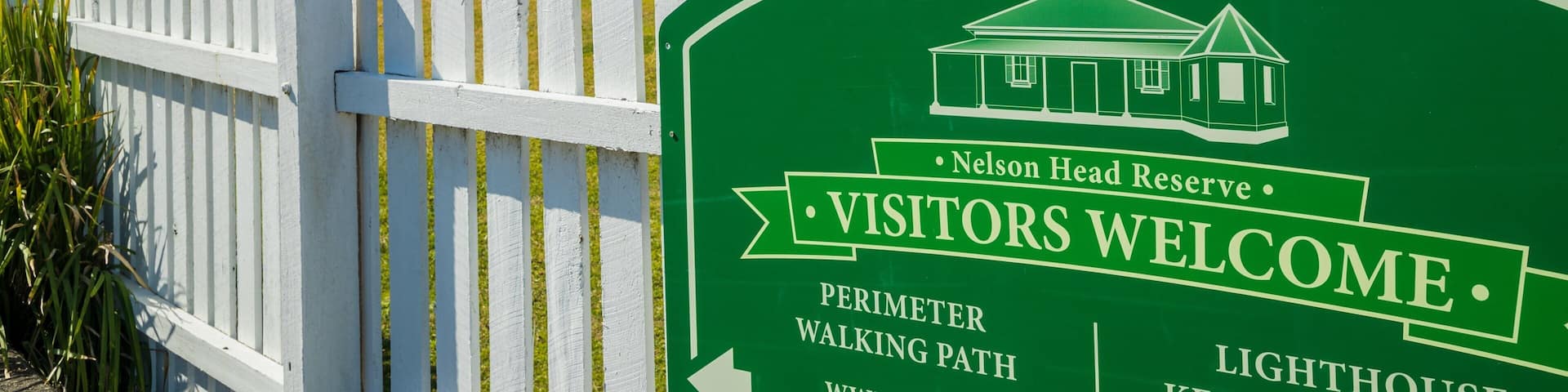 A green sign welcoming visitors to the Nelson Head Reserve, featuring a lighthouse and a perimeter walking path.