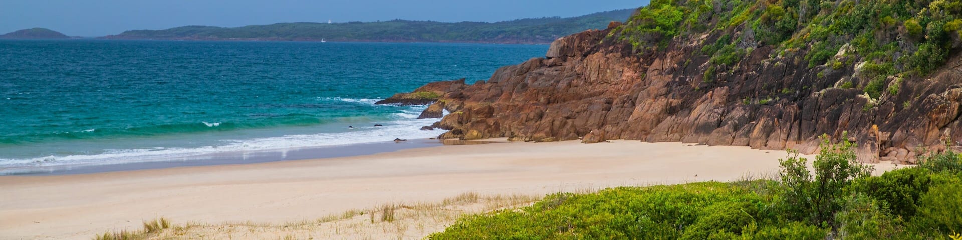 A beach with clear turquoise water, sandy shore, and a rocky cliff with greenery.