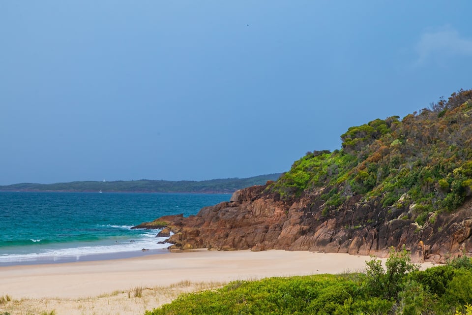 A beach with clear turquoise water, sandy shore, and a rocky cliff with greenery.