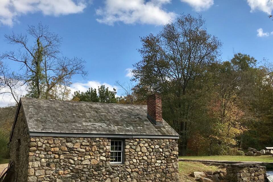 Una cabaña de piedra con chimenea, rodeada de árboles y un pequeño arroyo.