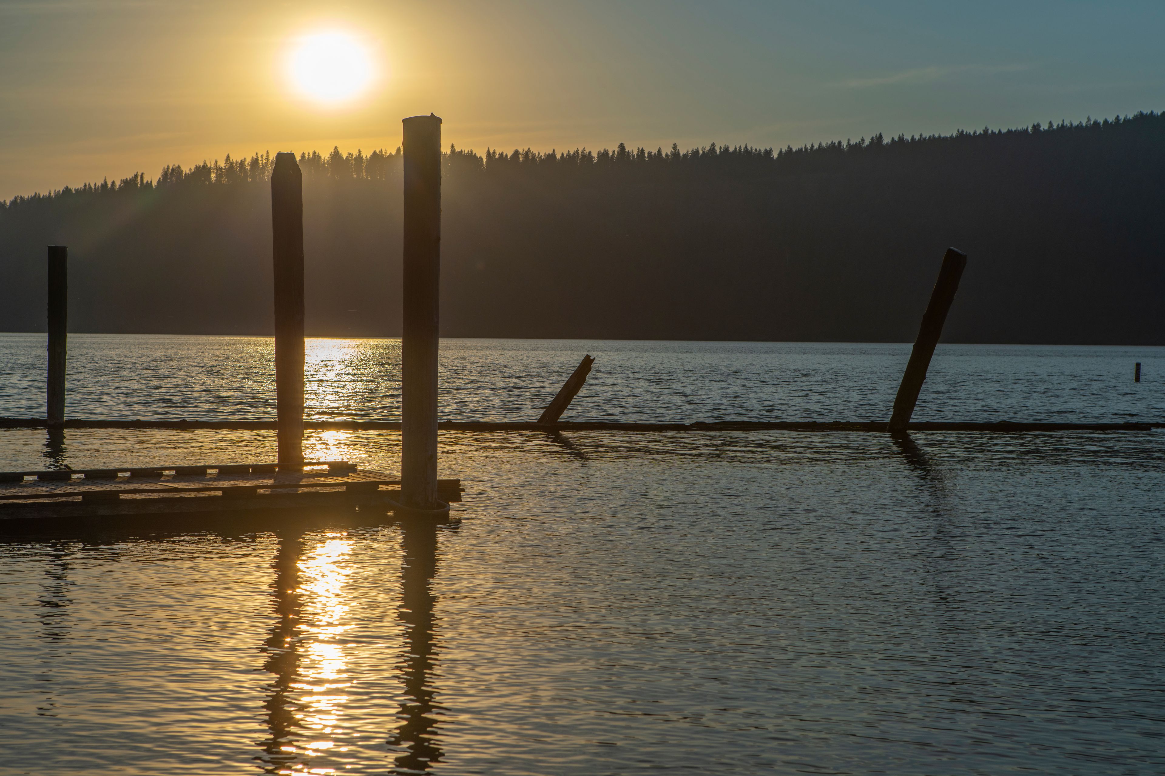 A sunset over a calm lake with a pier and a forested hill in the background.