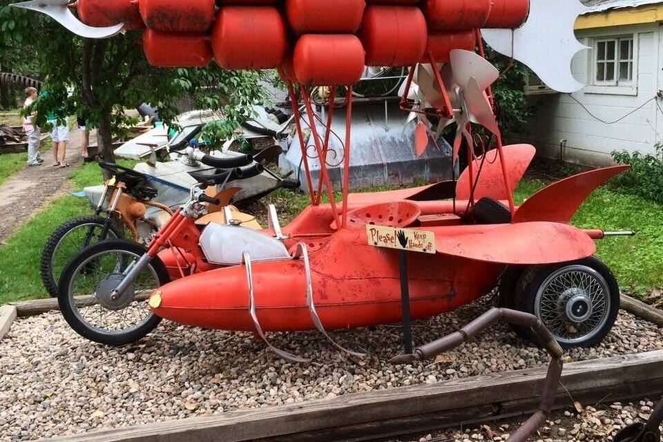 Um veículo vermelho peculiar com vários cilindros vermelhos numa estrada de cascalho, um letreiro com a inscrição "Please Keep Hand" e uma escultura de metal que se assemelha a um peixe.