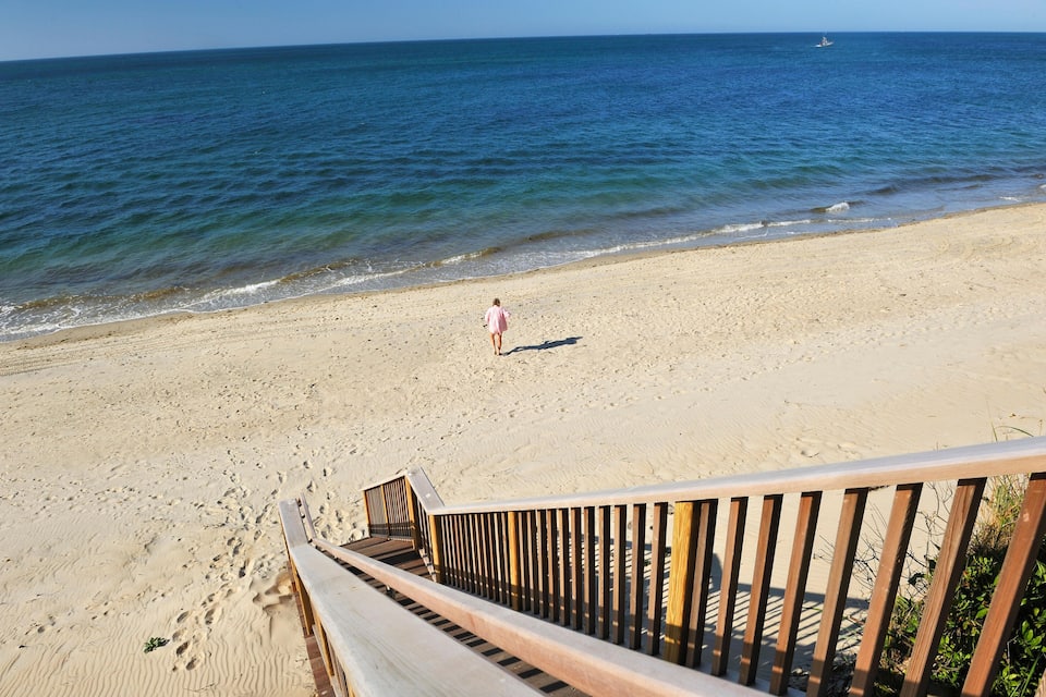 En strand med en trætrappe, der fører ned til sandet, en person går på stranden, og havet i baggrunden.