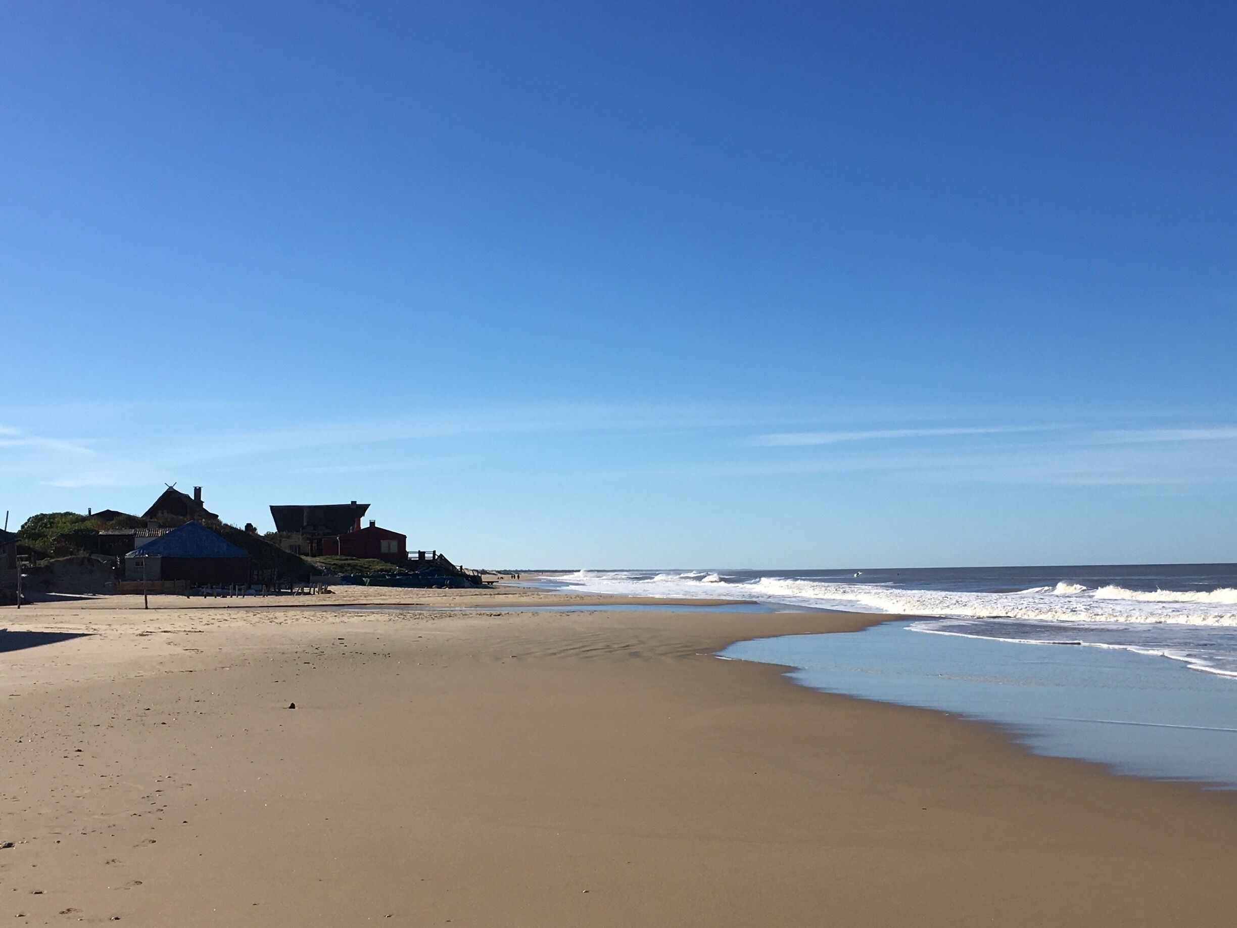 A sandy beach with waves, a clear blue sky, and a building with a blue roof.
