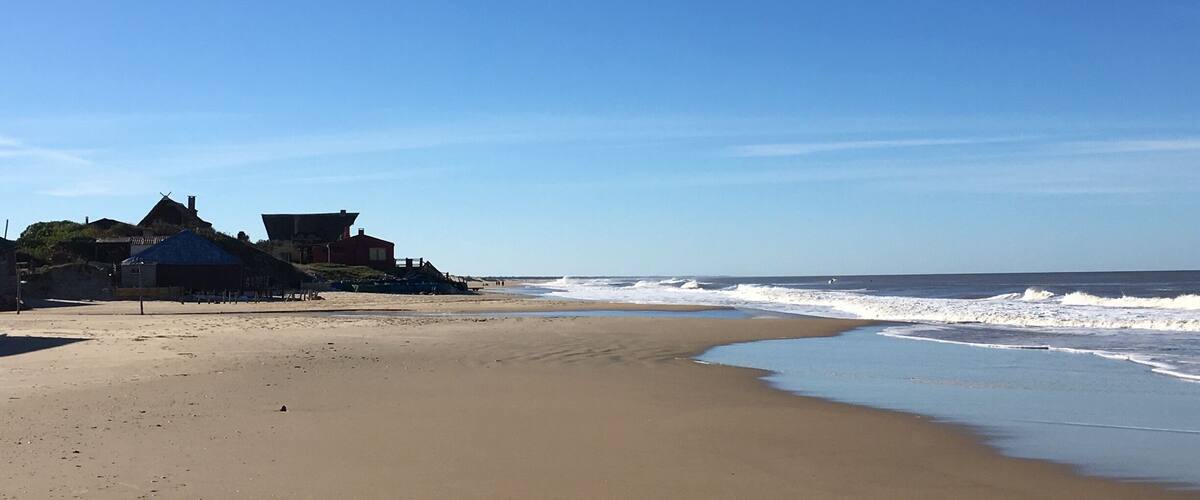 A sandy beach with waves, a clear blue sky, and a building with a blue roof.