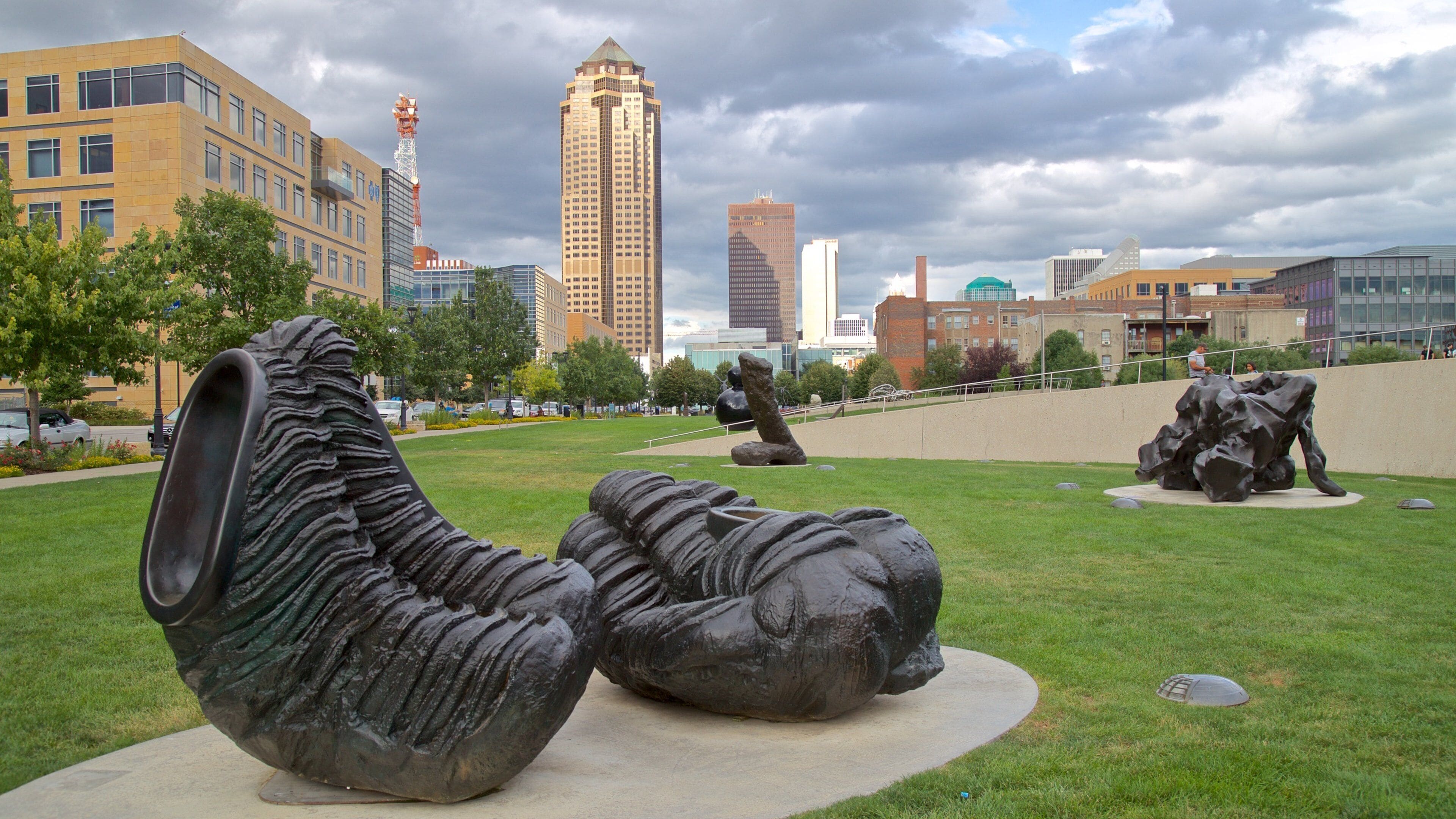 A sculpture garden with abstract metal sculptures in a grassy area, surrounded by modern buildings and a cloudy sky.