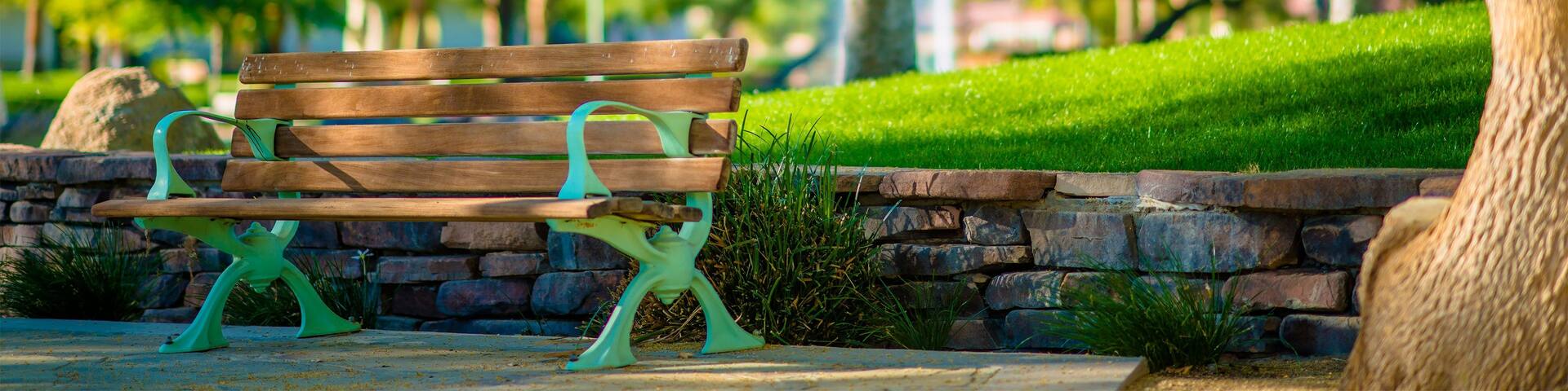 A park bench with a stone wall and grass, surrounded by trees and mountains in the background.
