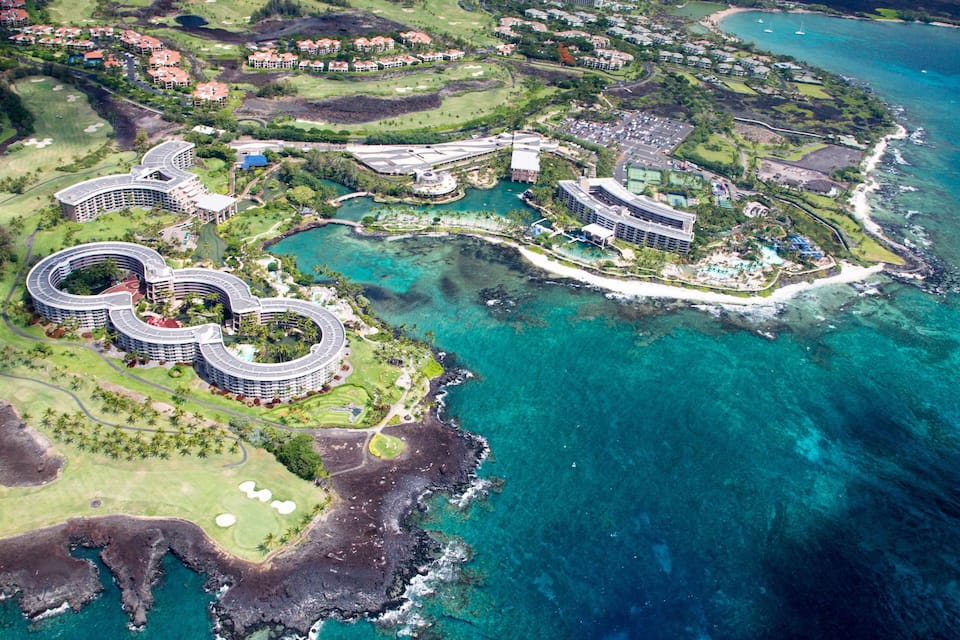 An aerial view of a coastal resort with multiple buildings, a golf course, and clear blue waters.