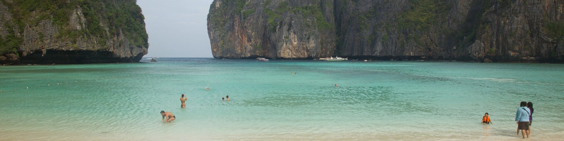 Uma praia com águas turquesa cristalinas, pessoas nadando e tomando sol, e imponentes falésias calcárias ao fundo.