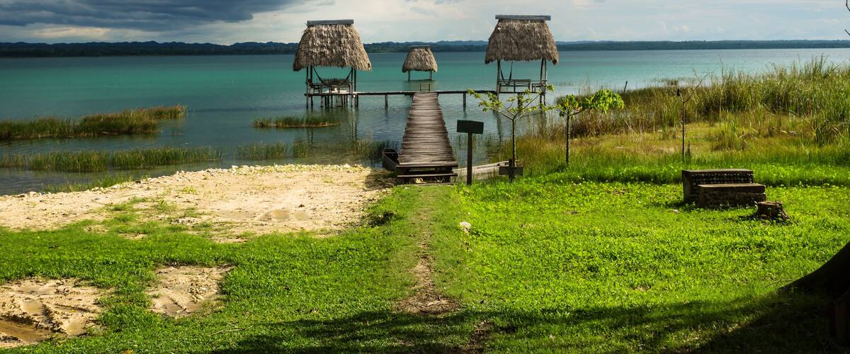 A wooden dock leading to thatched-roof huts by a calm lake with a cloudy sky.