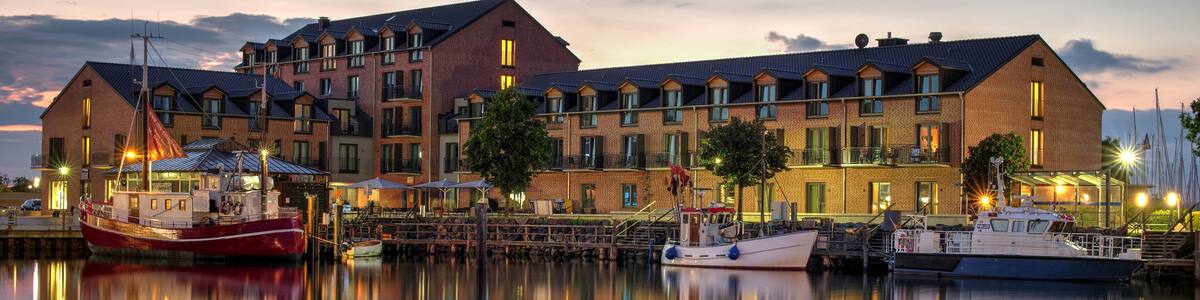 A waterfront building with multiple balconies, boats docked in the calm water, and a sunset sky.